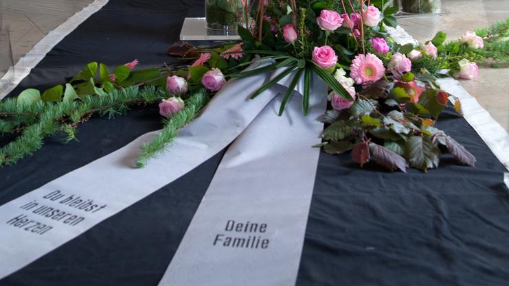 Blumenarrangement mit rosa Rosen und eine Trauerschleife vor einem Kreuz in einer Kapelle.