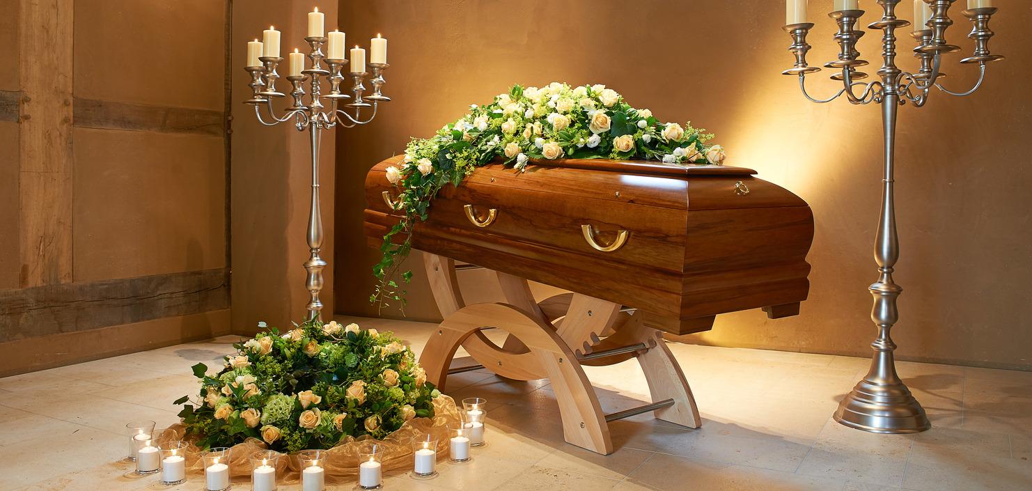 Sarg mit Blumenarrangement in einem Raum, flankiert von Kerzenhaltern und einer Blumendekoration auf dem Boden.