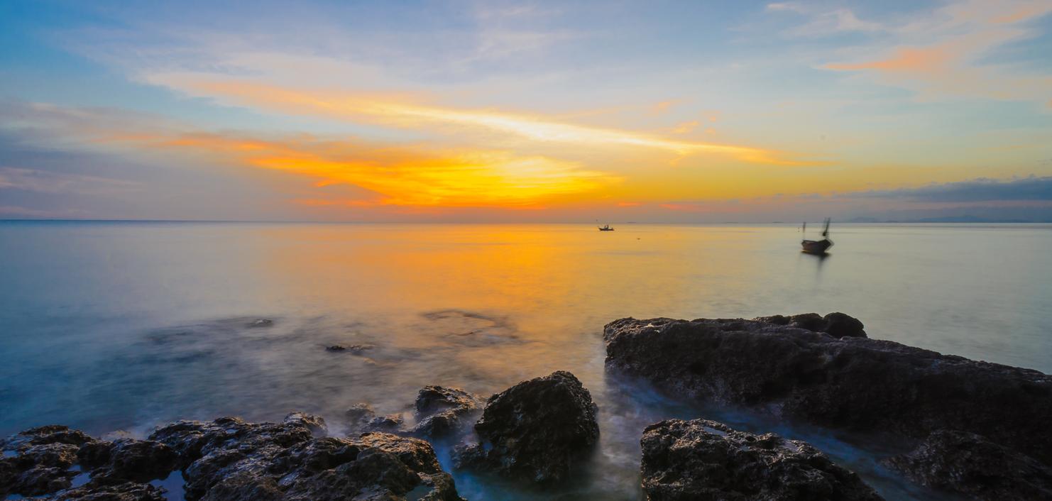 Sonnenuntergang über ruhigem Wasser, Felsen im Vordergrund und ein Boot entfernt auf dem Meer.