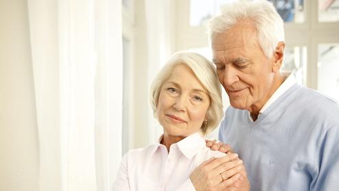 Ein älteres Paar steht nah beieinander, der Mann umarmt die Frau sanft, beide lächeln in eine ruhige Umgebung.